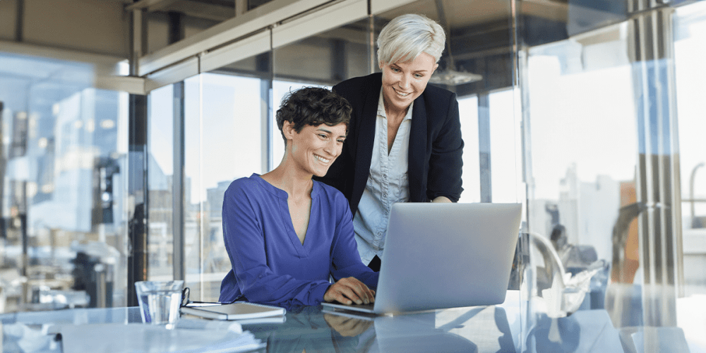 Two women, one standing and one sitting, are at a desk. They’re both looking at a laptop and smiling.
