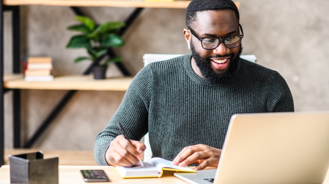 Man smiling and sitting at desk working on laptop and taking notes as he strategizes for providing CAAS