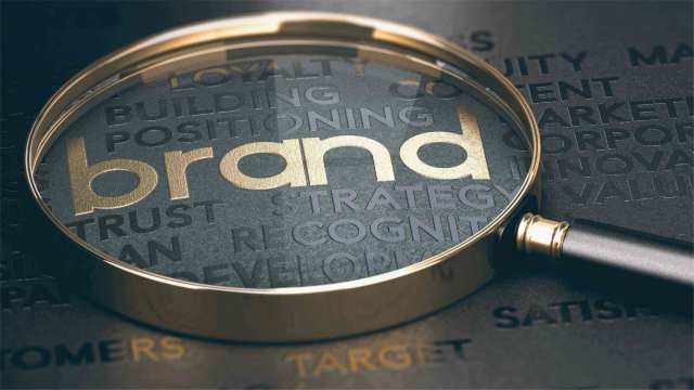 A word cloud sits on a black background. The word “brand” is in gold and is visible under a magnifying glass with a gold handle.