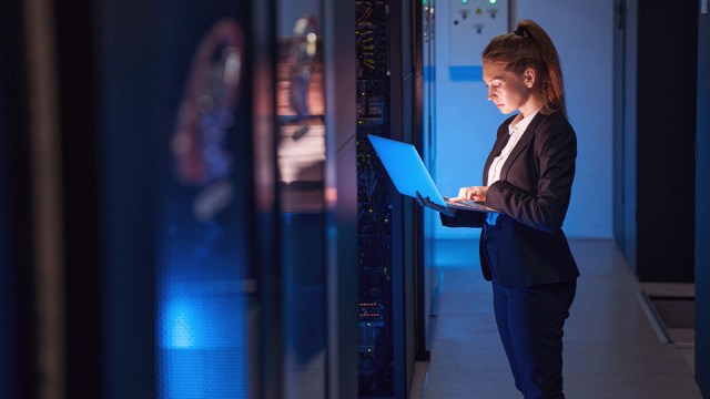 A woman stands in front of a server room, holding a laptop.