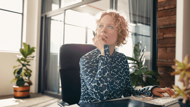 A woman with short, blonde, curly hair sits at her desk in an office. Her left hand sits poised on a stack of papers, while her right hand is tucked under her chin. She looks to the left, deep in thought as she contemplates cloud hosting services.