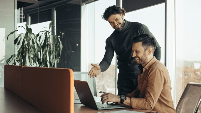 Two men, one seated and one standing, are all smiles while they look at a laptop screen and discuss adding cybersecurity to their list of client services.