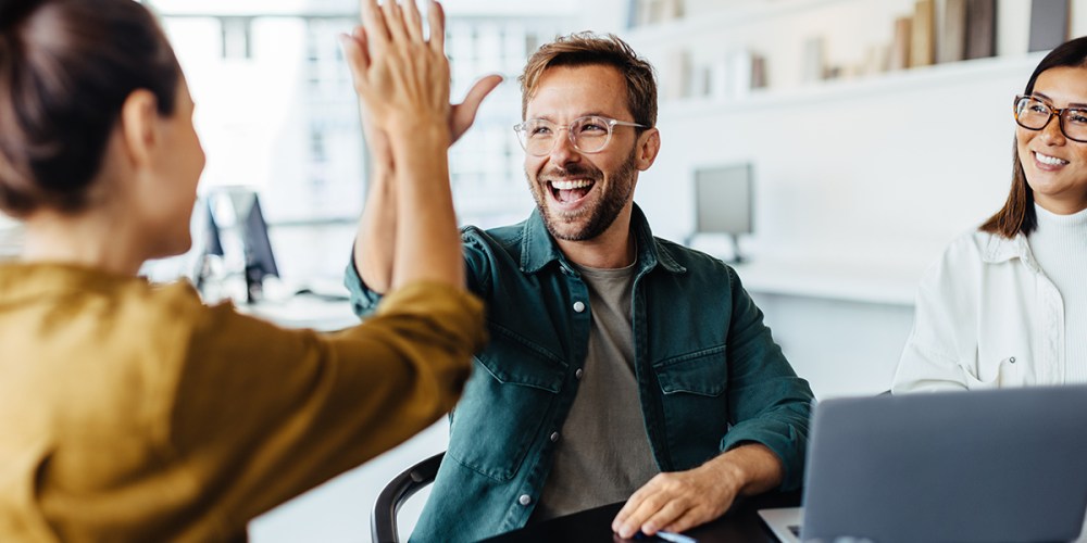 Three smiling people sit at a table in an office building. A man and a woman high five because they're on their way to building a healthier accounting firm.