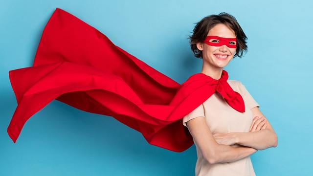 A woman with short brown hair stands on the right with her arms crossed and a smile on her face. She's wearing a red eye mask and a red flowing cape, looking like a superhero for being a Smart Team Manager.