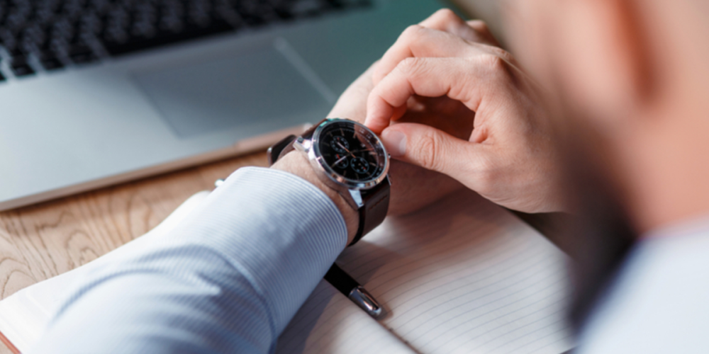 Image of a business person's arm and hand hovering over a desk with a laptop and notebook as they check their watch.