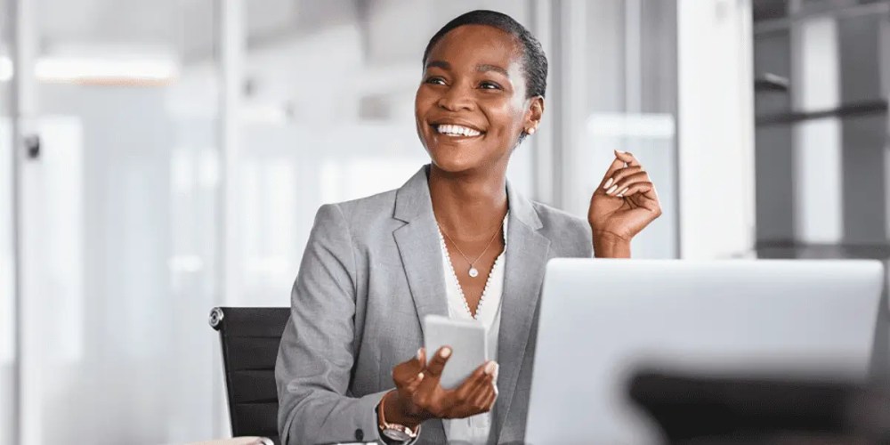 A smiling woman is sitting in front of a laptop with a smartphone in her hand.