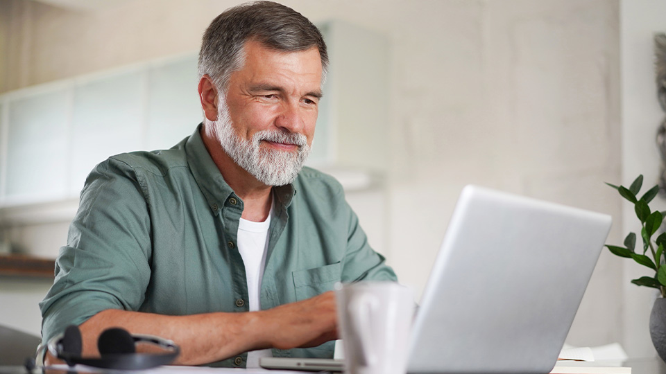 An older gentleman is smiling as he sits in front of a laptop.