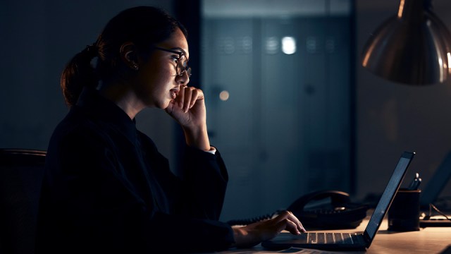 A woman sits in a darkened room, looking at a laptop.
