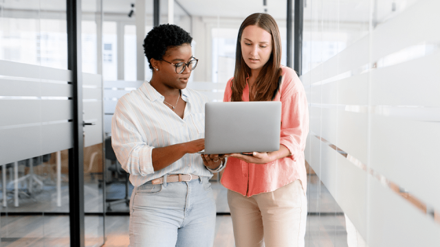 Two women stand while consulting over a laptop, one of them showing how AI has helped her jumpstart a client letter she needs to write.