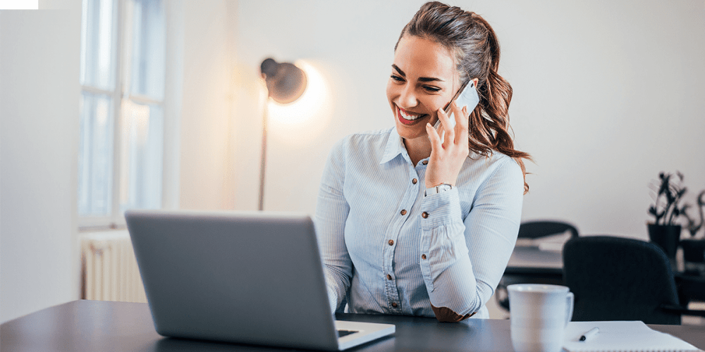 A woman sits smiling in front of a laptop, talking on the phone because AI in accounting just helped her complete a task more efficiently.