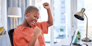 A female in an orange shirt sits at her desk smiling, with a laptop open in front of her while she has her hands in the air in celebration for the end of tax season.