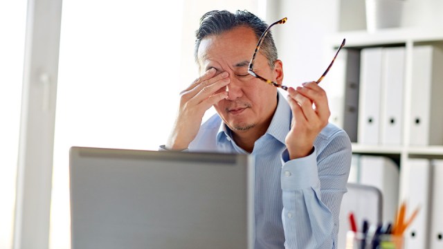 A man sits in front of a laptop, holding his glasses in one hand while rubbing his forehead with the other, eyes closed, looking tired and burned out after tax season.