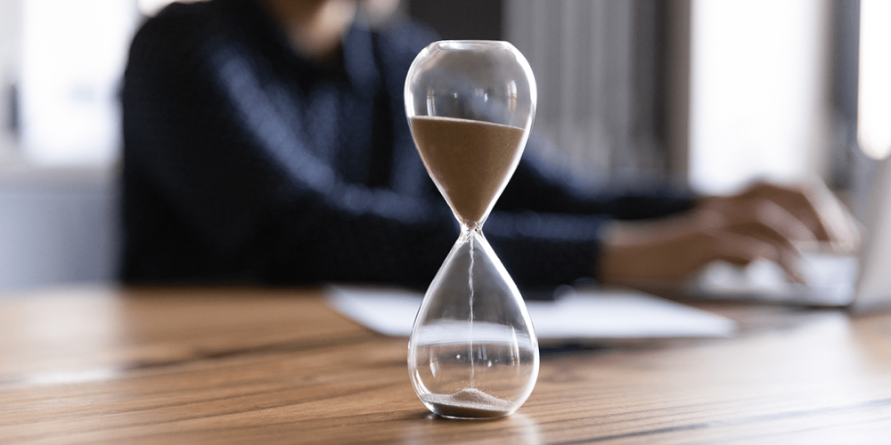 An hourglass sits on a desk in front of someone sitting at a laptop.