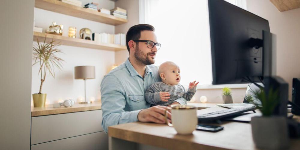 Man holding his baby in front of a computer while working remote