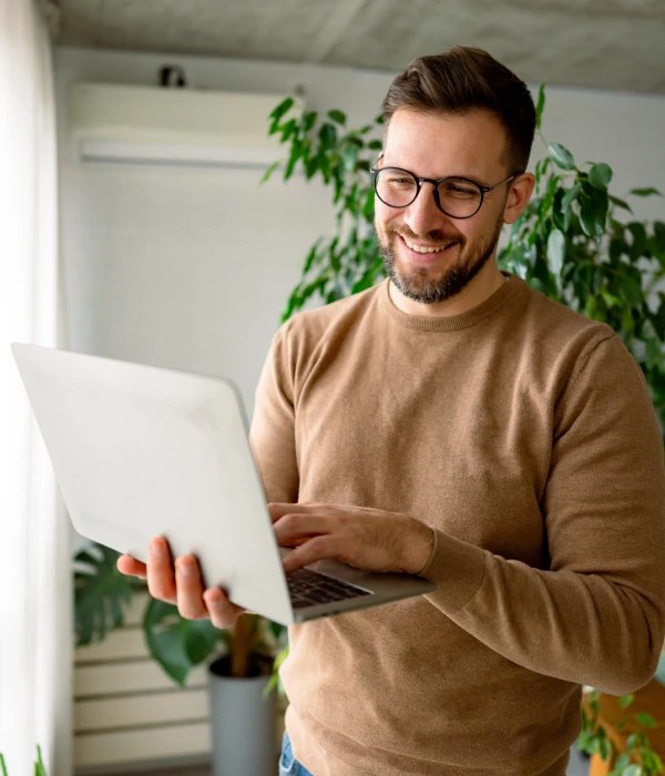 Person standing indoors using a laptop, with potted plants visible in the background.
