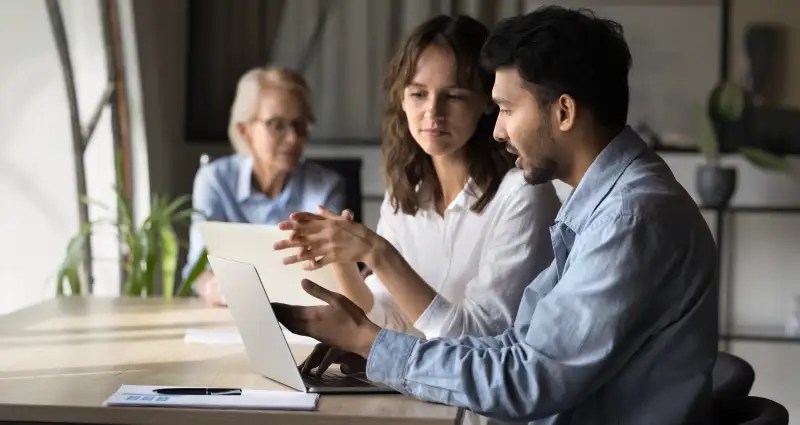 Two people in a discussion with a laptop screen and a third person in the background